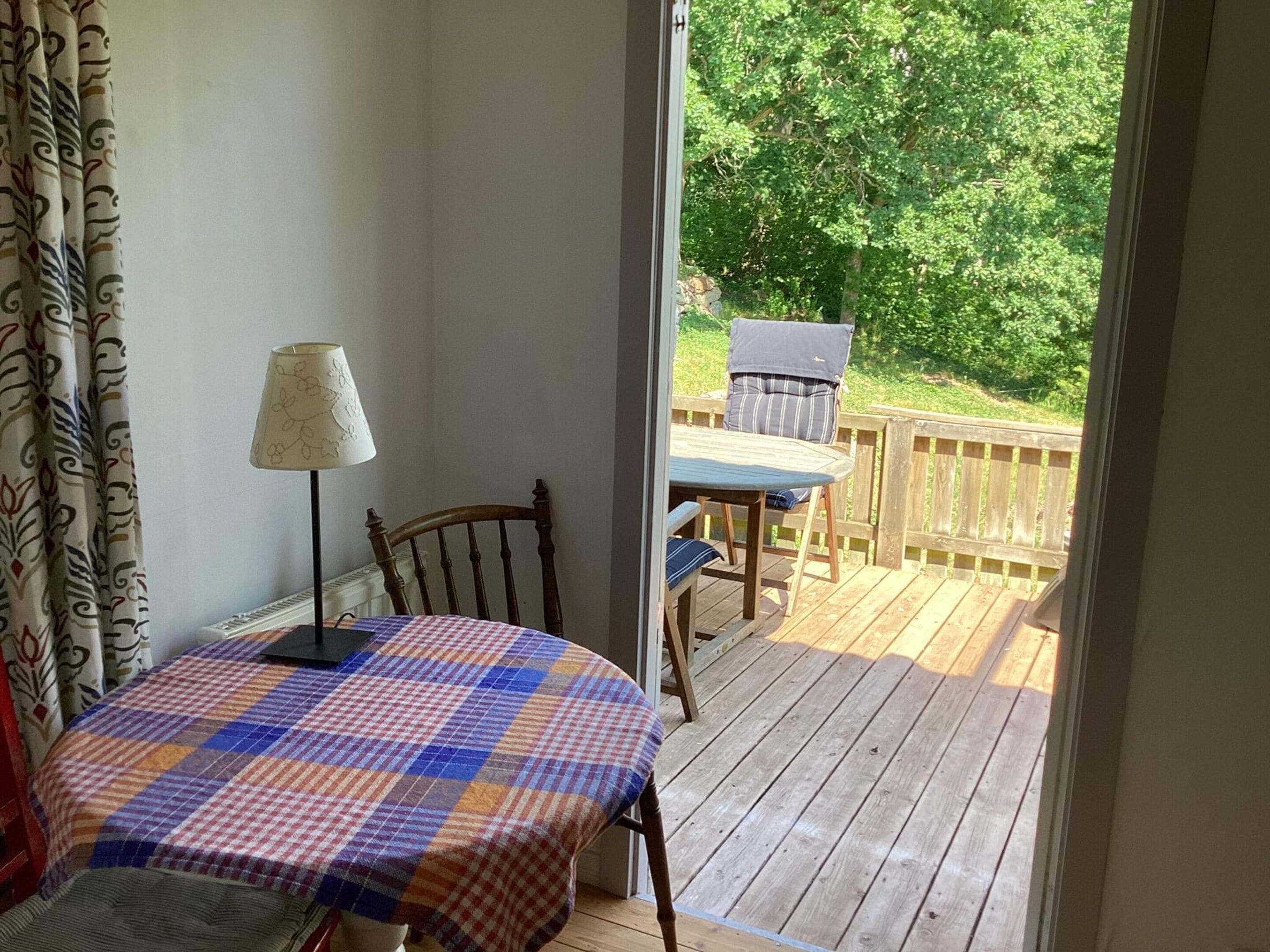 Una habitación con una mesa a cuadros, una lámpara y vistas a una terraza de madera y follaje exterior.