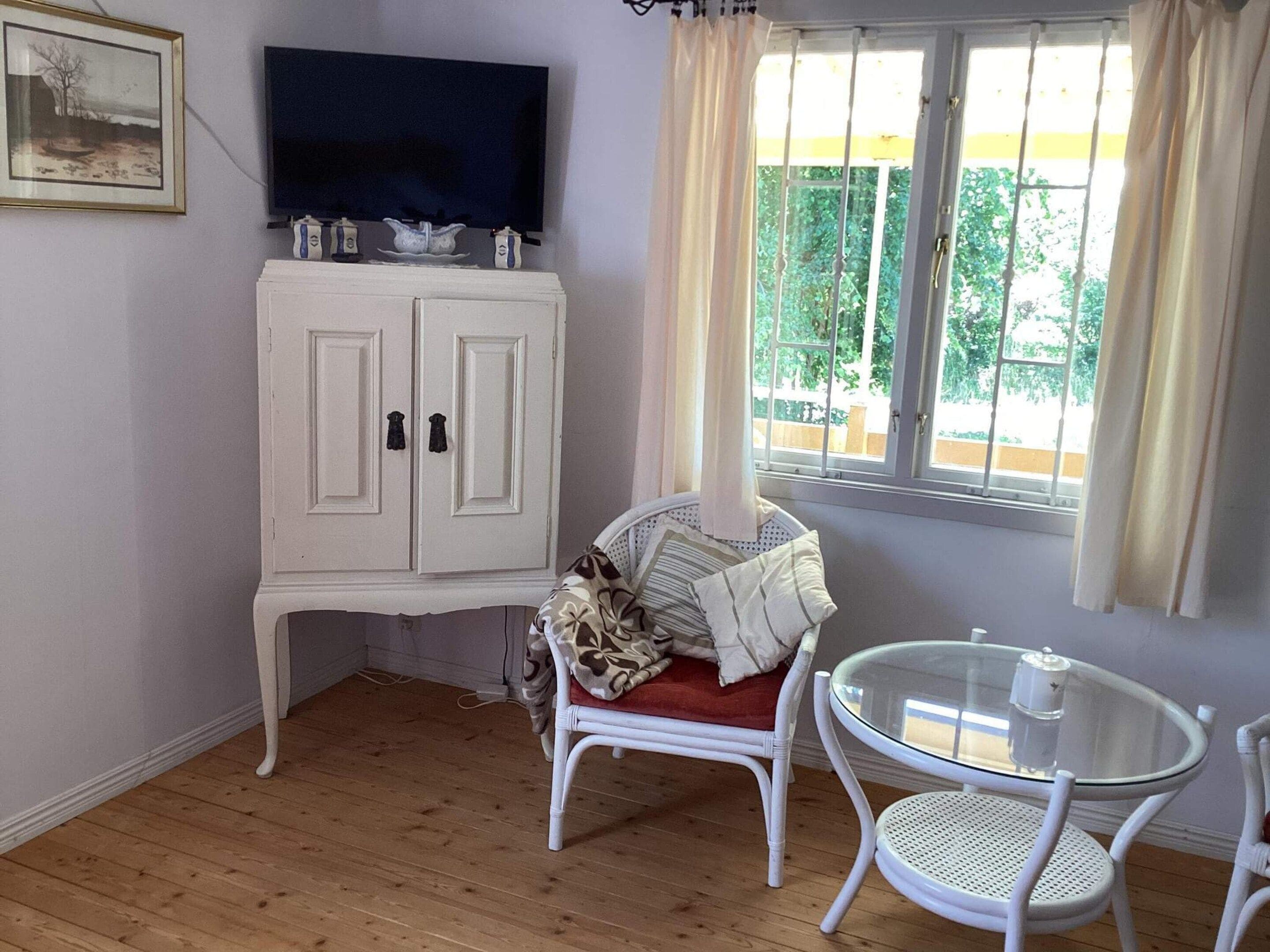 Una habitación con un armario blanco, un televisor, una silla de mimbre, una mesa de vidrio y una ventana con cortinas.