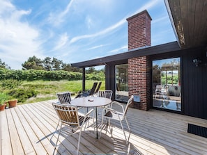 A wooden deck with a round table and chairs, a brick chimney, and a glass door leading to an outdoor area. - 6 Person Holiday Home in Henne (Henne)