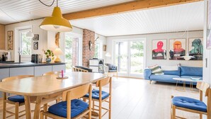A spacious living area with a wooden dining table, blue cushioned chairs, a blue sofa, and a kitchen area with a brick wall and a hanging light fixture. - 6 Person Holiday Home in Henne (Henne)