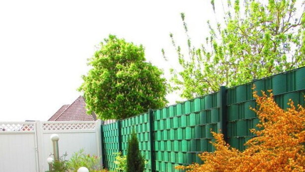 Ein Garten mit einem gefliesten Weg, einem grünen Zaun und verschiedenen Sträuchern und Bäumen.