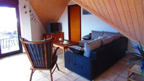 Ein gemütliches Loft mit Holzbalkendecke, einem dunkelblauen Sofa, einem Holzstuhl, einem kleinen Tisch und einem Fernseher auf einem Holzschrank.