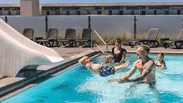 Una piscina con tobogán, sillas de descanso y un edificio al fondo.