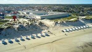 A coastal hotel with multiple buildings, a sandy beach with beach huts, and a forested background.
