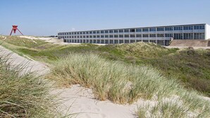 Um edifício moderno com telhado plano e amplas janelas, rodeado por dunas de areia e relva.