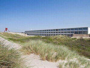 Un edificio moderno con techo plano y ventanas amplias, rodeado de dunas de arena y hierba.