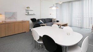 Quarto moderno e bem iluminado com mesa de jantar branca, cadeiras pretas, sofá cinza e armário de madeira.