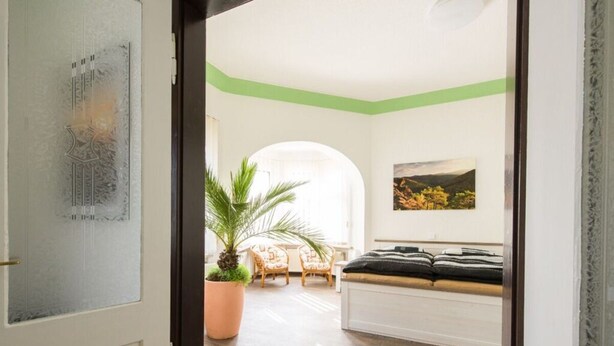 A room with a wooden floor, a bed with black bedding, a large potted plant, and a painting on the wall. - Holiday Apartment Angela Koenig (Wernigerode)