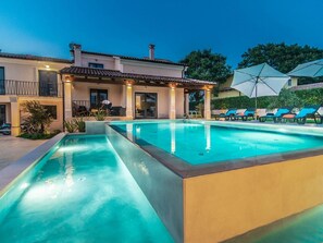 A rectangular swimming pool with a tiled deck, a covered patio area with outdoor seating, and a two-story building with balconies.