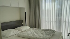A neatly made bed with a quilted blanket and pillows, a wooden headboard, and a view of a cityscape through sheer curtains. - Apartment in Nieuwpoort for 6 Persons (Nieuwpoort)