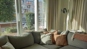 A modern living room with a sectional sofa, a floor lamp, and a view of the street through large windows. - Apartment in Nieuwpoort for 6 Persons (Nieuwpoort)
