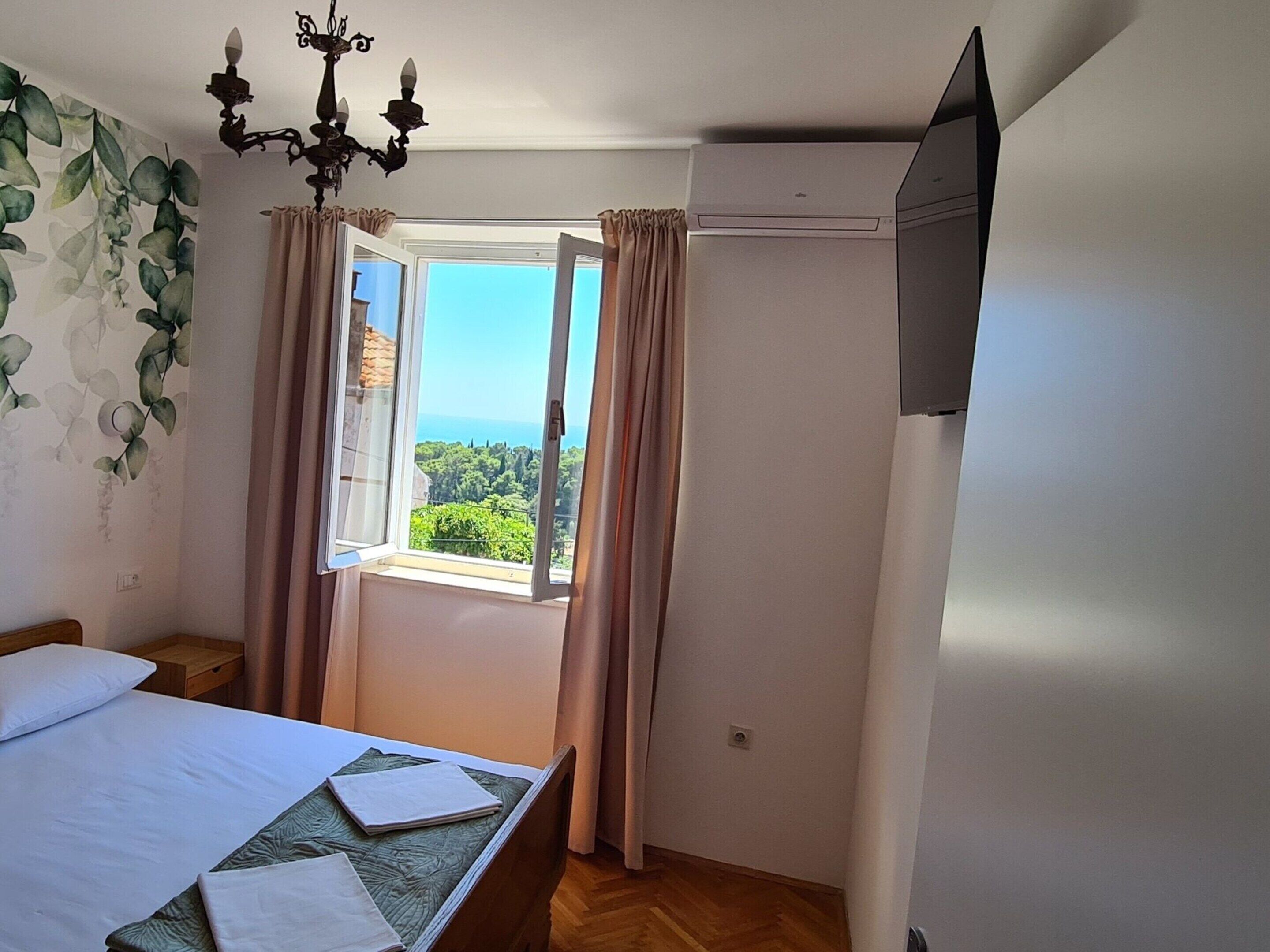 A bedroom with a bed, a window with curtains, a chandelier, and a view of greenery outside.