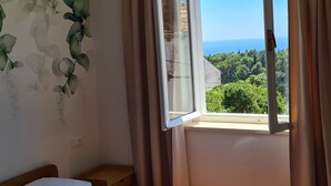 A room with a bed, a small wooden table, a window with curtains, and a view of greenery and the sea. - Apartment Solvit - Three Bedroom Apartment With Terrace (Dubrovnik)