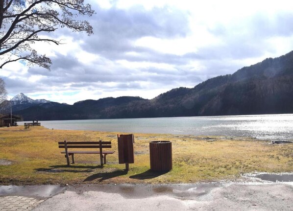 A lakeside park with a bench and trash can, surrounded by mountains and a cloudy sky. - Ferienhaus im Allgau Spass und Naturaufenthalt (Nesselwang)