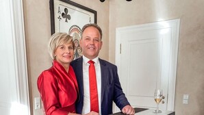 A man and a woman standing in a room with a framed playing card on the wall, a white door, and a bar area with a glass of wine. - Villa _master Suite Comfortable Vacation Home (Ilsenburg)
