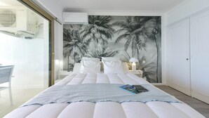 Een slaapkamer met een groot bed, wit beddengoed, een muurschildering van een palmboom, een nachtkastje met een lamp en een stoel in de hoek.