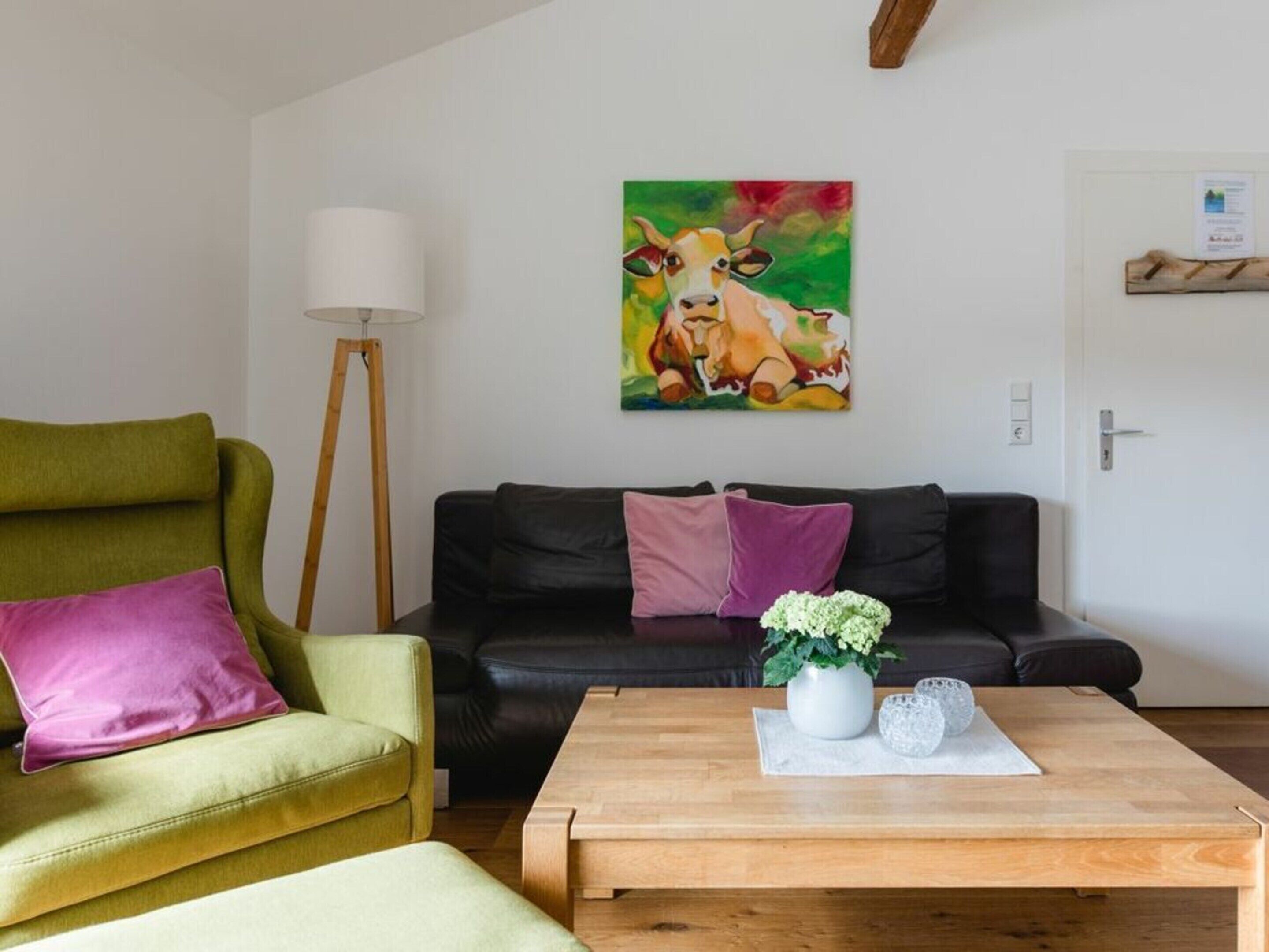 Un salon avec un fauteuil vert, un canapé en cuir noir et une table basse en bois. Il y a un tableau représentant une vache accroché au mur et une lampe sur pied.