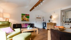 A living room with a sofa, armchair, and ottoman, a coffee table with decorative items, a painting on the wall, and a kitchen area with a microwave and fruit bowl. - Krahenberg Country House (Eslohe)