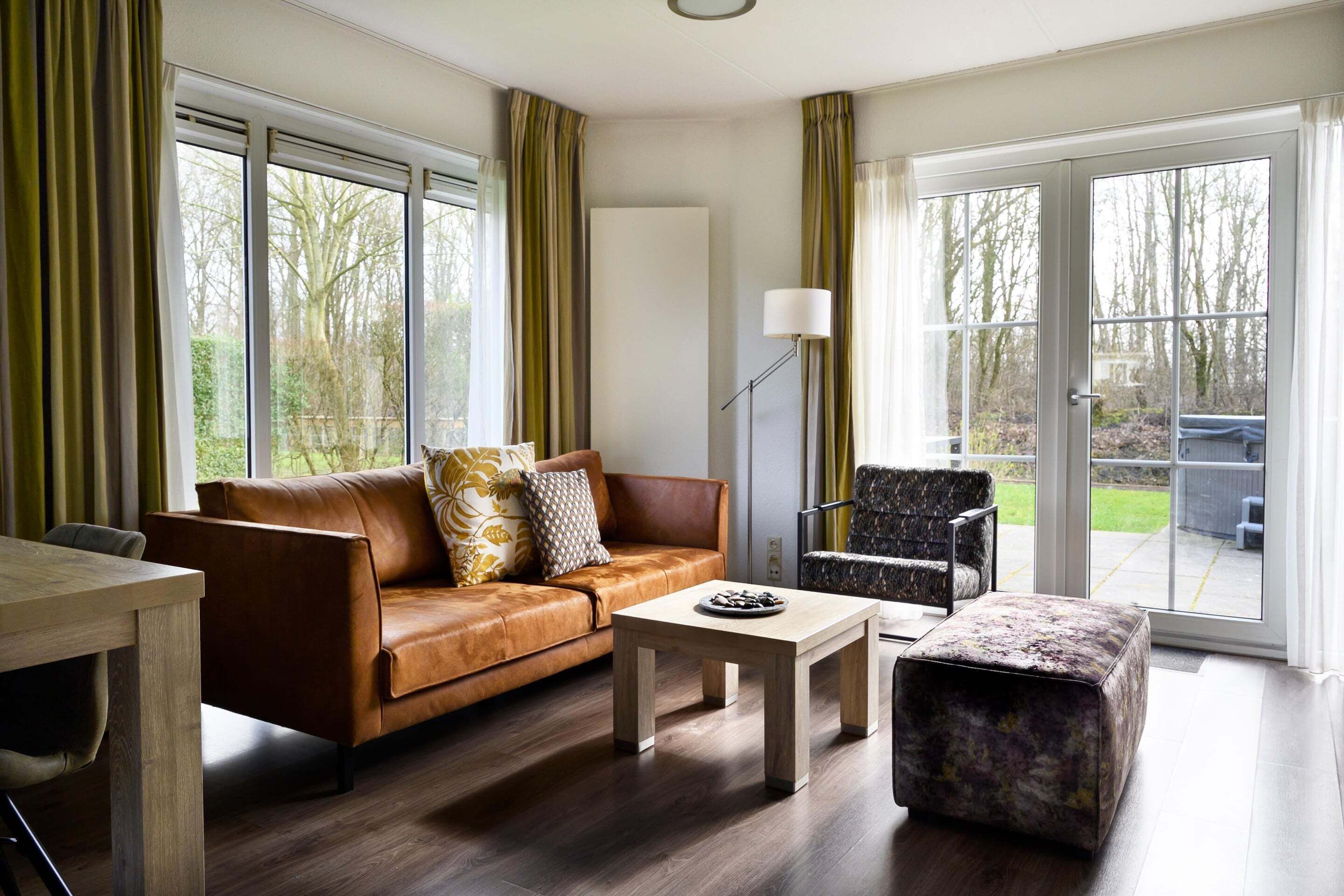 A modern living room with a leather sofa, a coffee table, a patterned ottoman, a floor lamp, and large windows with curtains.