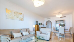 A modern living room with a beige sofa, a glass coffee table, and a dining area with a white table and chairs. - Belvilla by OYO La Zenaida II (Rincon de la Victoria)