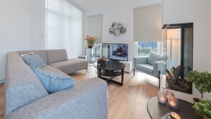 Ein modernes Wohnzimmer mit einem grauen Sofa, einem kleineren grauen Sessel, einem Couchtisch und einem Flachbildfernseher. Rechts befindet sich ein Kamin mit einem Baum und einer Pflanze, links ein Fenster mit Jalousien.
