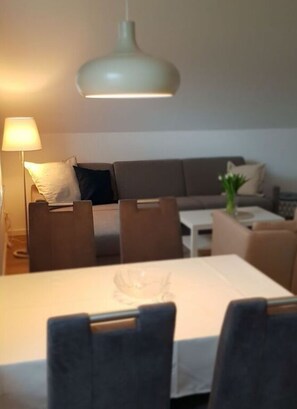 A dining area with a table set for two, a sofa, and a lamp in the background. - Holiday Apartment Bathmann (Cuxhaven)