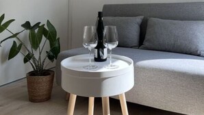 A modern living room with a grey sofa, a round white side table with two wine glasses and a bottle, and a potted plant. - Sartoris Comfortable Holiday Residence (Bad Neuenahr-Ahrweiler)