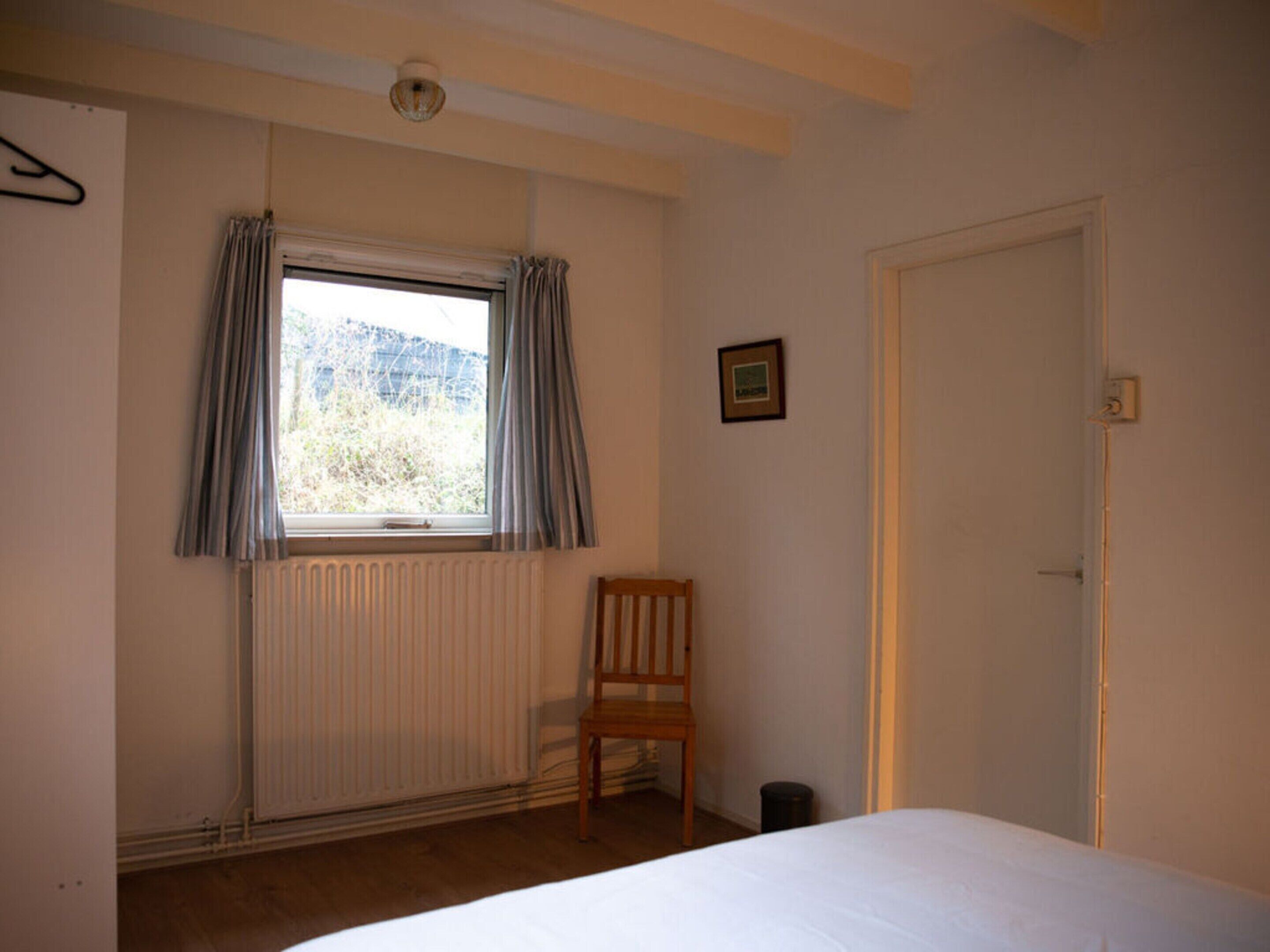A bedroom with a single bed, a radiator, a window with curtains, a wooden chair, and a door.