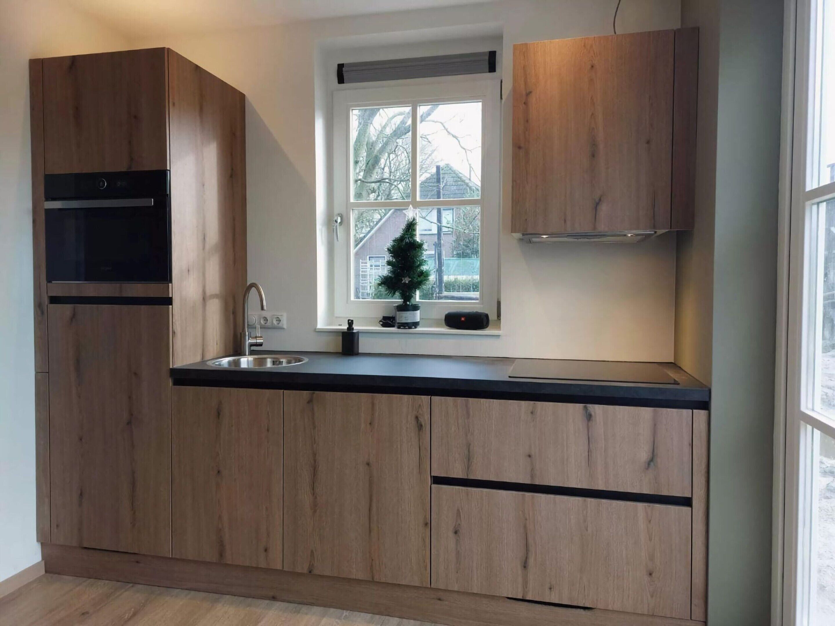 A modern kitchen with wooden cabinets, a built-in oven, a stainless steel sink, and a window with a view of trees and a house.