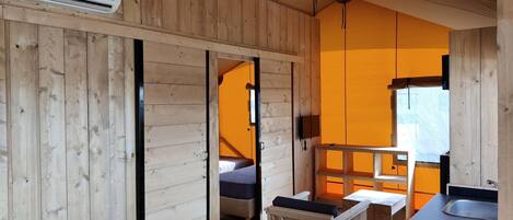 Interior de una cabaña de madera con una cama, una mesita, un lavamanos y un aire acondicionado.