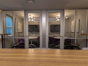 A conference room with a wooden table, two purple chairs, a ceiling light fixture, and a window with blinds.