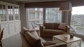 Una sala de estar con un sofá, una mesa de centro y un aparador. Se ve una ventana con vistas a edificios.