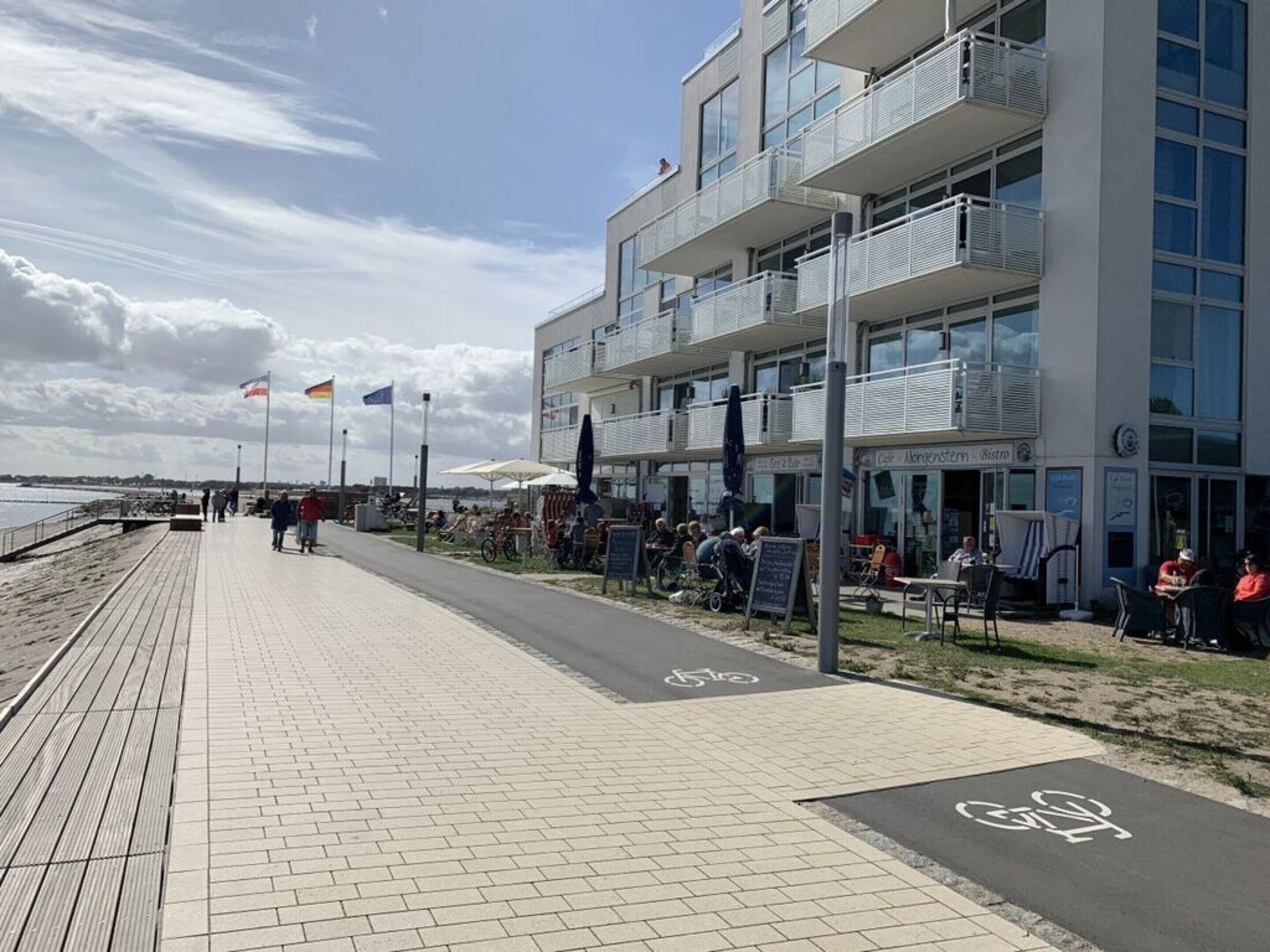 A waterfront promenade with a bike lane, outdoor seating, and a modern building with balconies.