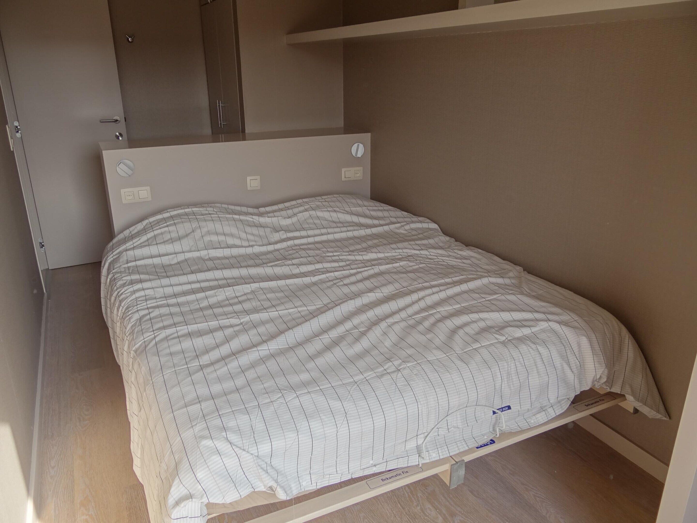 A single bed with a white striped bedspread in a room with a wooden floor and a beige wall.