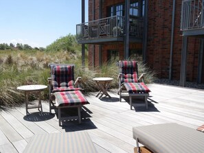 A deck with two lounge chairs and a small table, overlooking a grassy area and a brick building with a balcony.