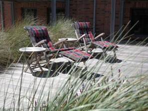A patio area with two lounge chairs and a small table, surrounded by tall grasses and a brick building in the background.