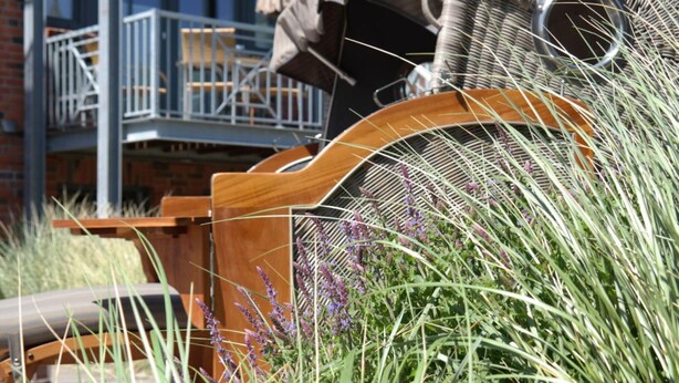 A wooden bench with a woven basket and a metal grill, surrounded by tall grass and purple flowers. - Captain House Wangerooge - Nautik Suite (Wangerooge)