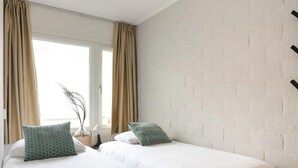 Habitació d'hotel amb dos llits, cortines beix, paret de maó blanc i una finestra amb cortines.