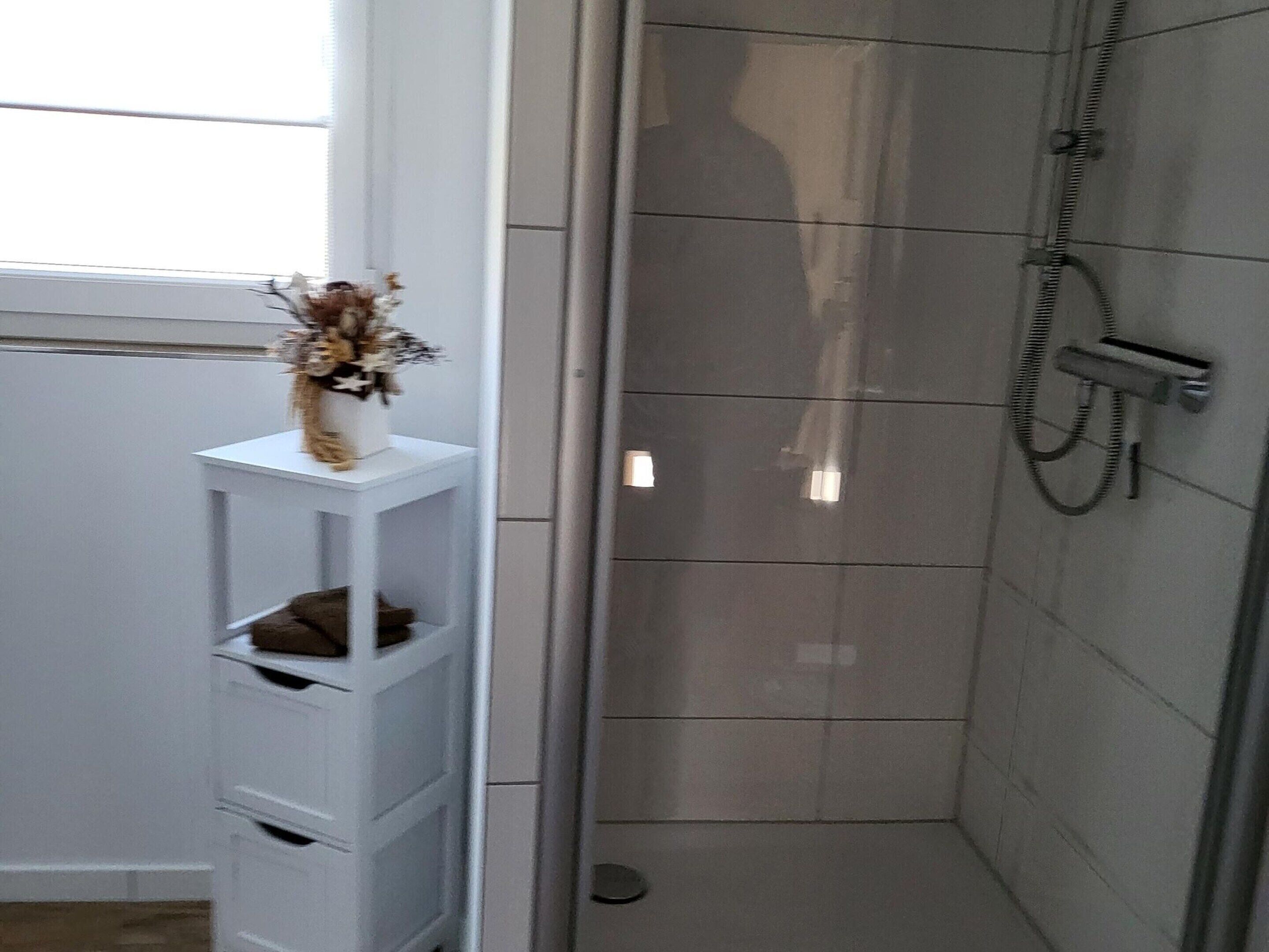 A bathroom with a shower, a white shelf with a vase, and a window.
