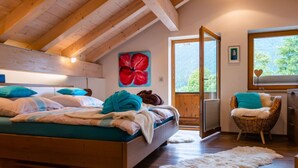 A bedroom with a wooden ceiling, a bed with teal bedding, a large window, and a wicker chair. - Apartment in Garmisch-partenkirchen (Garmisch-Partenkirchen)