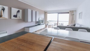 A modern living room with a wooden dining table, a sofa, and a TV on a white cabinet. There are large windows with curtains and a view of the outside. - Apartment Nieuwpoort Near Beach Promenade (Koksijde)