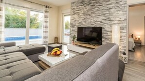 Un salon moderne avec un mur en pierre, un téléviseur à écran plat, des canapés gris et une table basse sur laquelle sont disposés des fruits.