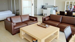 Un salon moderne avec deux canapés en cuir marron, une table basse en bois et, au fond, un coin cuisine.