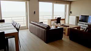 Un salon moderne avec un canapé, une table à manger et un téléviseur. De grandes fenêtres offrent une vue sur l’océan.