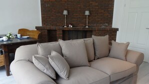 A beige sofa with multiple cushions in a living room with a brick fireplace and a white door in the background. - Anchorage Modern Retreat (Kappeln)