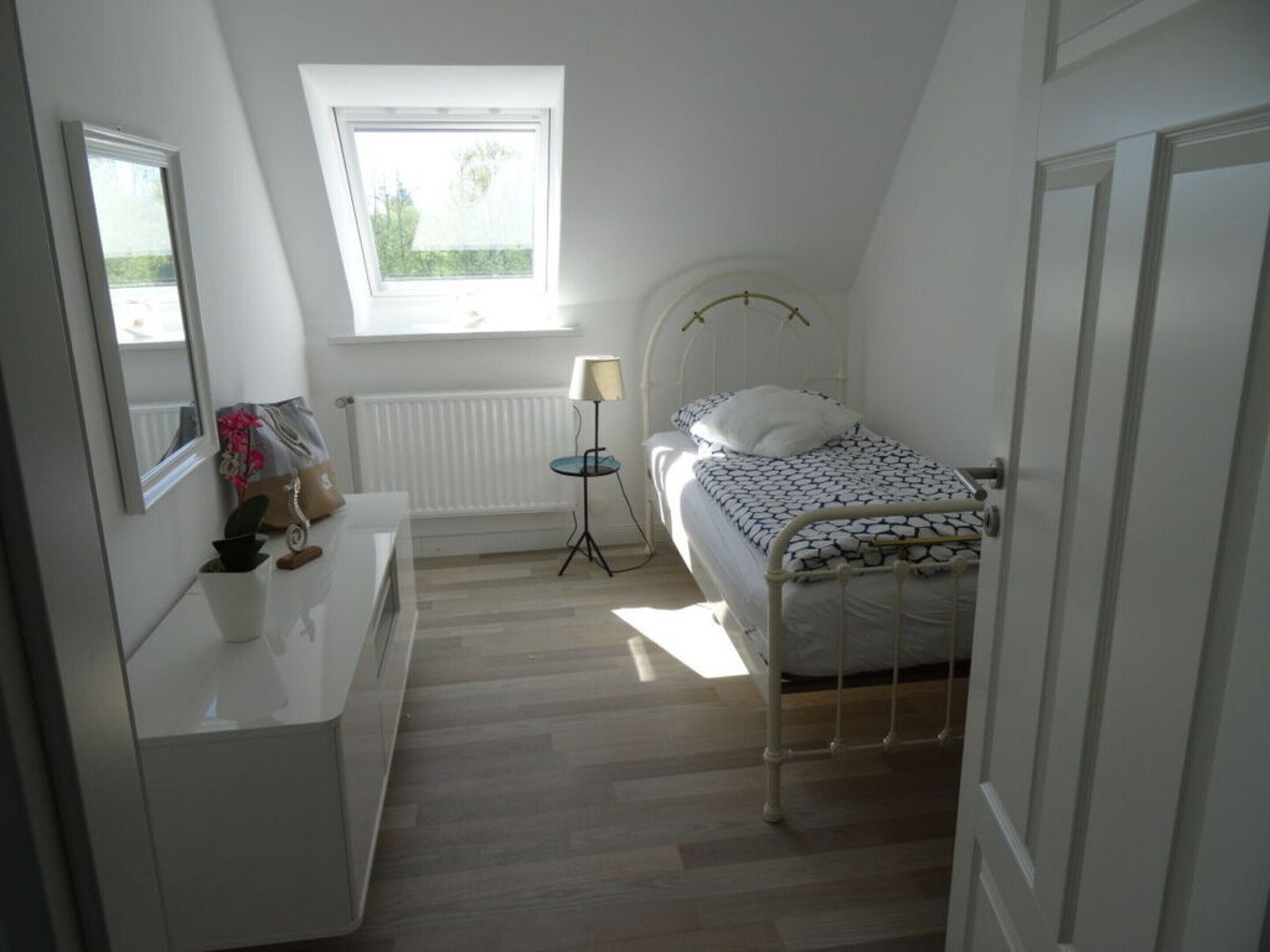A small, well-lit bedroom with a single bed, a bedside table, a lamp, and a window with a view of greenery.