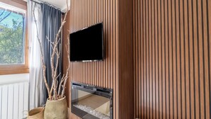 Una sala de estar moderna con una pared de paneles de madera, un televisor de pantalla plana sobre la chimenea, un ventanal con cortinas y una planta en maceta.