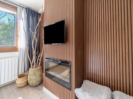 Una sala de estar moderna con una pared de paneles de madera, un televisor de pantalla plana sobre la chimenea, un ventanal con cortinas y una planta en maceta.