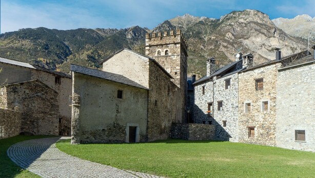 Un edificio de piedra con una torre, rodeado por un sendero adoquinado y una zona verde, con montañas al fondo.