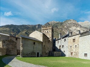 A stone building with a tower, surrounded by a cobblestone path and grassy area, set against a mountainous backdrop.